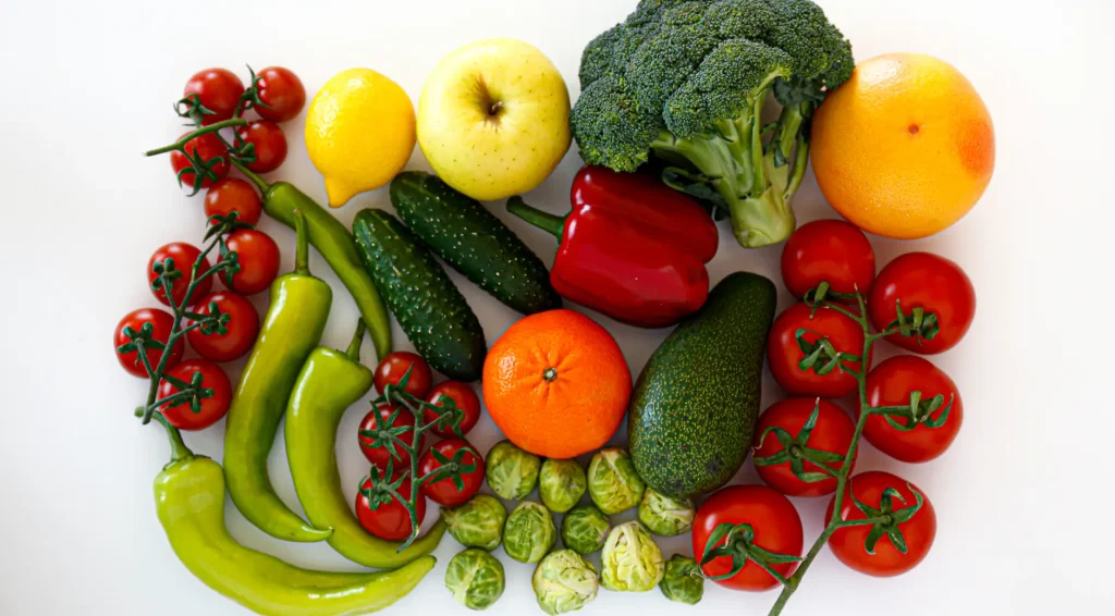 Clean, ready-to-package assortment of fresh vegetables and fruits including tomatoes, peppers, broccoli, bananas, and citrus on white background